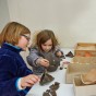 An einem Tisch arbeiten Kinder. Sie setzen Tonscherben zu Gefäßen zusammen. Auf der linken Seite sitzen zwei Mädchen, die gemeinsam ein braunes Gefäß zusammenpuzzeln. Auf der rechten Seite beugt sich gerade ein Junge über ein halb fertiges weißes Gefäß, während er Teile mit Kreppklebeband zusammenklebt. 