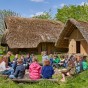 Im Freien sitzt eine Schulklasse im Kreis. Im Hintergrund befinden sich zwei Lehmhäuser mit Strohdach: ein großes Wohnhaus und rechts davor ein kleineres Gebäude.