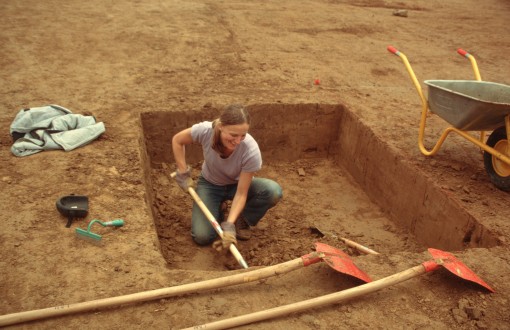Eine Frau kniet in einem rechteckigen Erdloch und gräbt. Im Vordergrund liegen zwei Schaufeln und links steht eine Schubkarre.