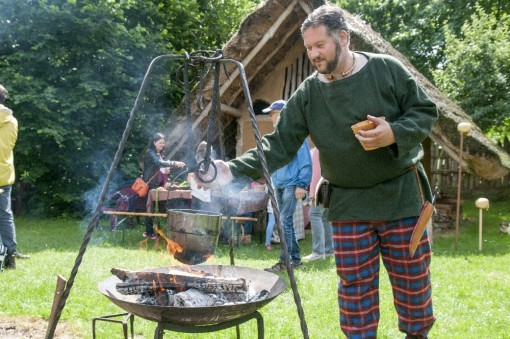 Foto: Ein Aktuer in keltischem Gewand steht an einem Lagerfeuer. Über dem Feuer befindet sich ein Dreibein mit Topf, imHintergrund Personen und ein Lehmhaus.