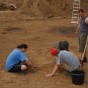 Foto: Grabungssituation, im Vordergrund hocken Grabungsmitarbeiter und legen mit Kellen Befunde im Erdreich frei. Im Hintergrund, auf einem Hügel stehen zahlreiche Besucher.