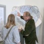 Foto: Auf dem Bild zeigt ein Mitarbeiter einer Besucherin auf ein Ausstellungsstück an der Wand. an der Wand hängen Poster. 