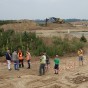 Im Vordergrund liegen mehrere Grabkammern, mit Feldsteinen ausgekleidet oder ohne Grabeinfassung, die archäologisch freigelegt wurden. Im Mittelgrund befindet sich ein Weg, der mit einer Absperrung von der Grabungsfläche abgegrenzt ist. Hier unterhalten sich Archäologen über die Begrenzung hinweg mit den Besuchern. Im Hintergrund steht ein Bagger auf einem Erdhügel.