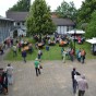 Bild des Innenhofes. Links und im Hintergrund sind Gebäudeteile erkennbar. Auf der Wiese sitzen zahlreiche Besucher an Tischen und dort stehen zwei Verkaufswagen. Im Vordergrund laufen und stehen Besucher auf der gepflasterten Einfahrt.