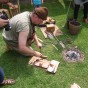 Der Bronzegießer kniet auf der Wiese. Vor ihm befindet sich ein Glutbett aus Kohlen und Geräte für den Bronzeguss. Um ihn herum sieht man die Beine mehrerer Zuschauer.