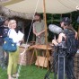 Ein dreiköpfiges Kamerateam steht vor einem Händlerstand. Zwei Personen tragen Kameras auf ihren Schultern. Der historisch gewandete Händler steht hinter seinem Tisch voller Waren, der mit einer Zeltplane überspannt ist.