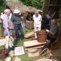 Ein Korbflechter sitzt vor einem Holzhaus mit Strohdach. Vor ihm stehen einige Besucher und einige Körbe. Einen großer Korb mit 2 Henkeln bearbeitet er gerade. Im Hintergrund befindet sich ein Marktstand.