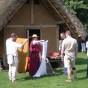 Vor einem Lehmhaus mit Strohdach stehen Beuscher und zwei Personen in historischem Gewand. Sie trägt ein rotes Kleid und einen Stohhut und hält ein weißes Tuch in den Händen. Er trägt ein hellbeiges Gewand aus Hose und Hemd mit Messer am Gürtel.