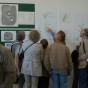 Besucher im Ausstellungsraum. Im Hintergrund Poster mit Abbildungen der Holzteile der Wassermühle.