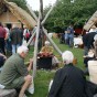 Eine historisch gewandete Färberin sitzt am Feuer neben ihrem Kessel. Um sie herum sieht man Besucher und Gebäude der eisenzeitlichen Hofanlage.