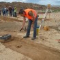 Ein Kollege der Grabungsmannschaft hält das Erdreich mit einer Wasserspritze feucht, hinter ihm stehen ein Stativ mit Nivelliergerät und Besucher.