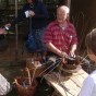 Zwei Korbflechter sitzen vor einem Haus, einer arbeitet an einem Weidenkorb. Im Vordergrund befinden sich Besucher mit Kindern und zahlreiche Korbwaren.