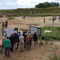 Blick über die Grabungsfläche mit Besuchern vor Informationstafeln, mit Flatterband gekennzeichneten Wegen und rechts im Bild einer Teilrekonstruktion eines Hauses aus Holz.