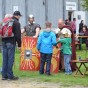 Bild vom Innenhof der Außenstelle Titz. Zwei römische Legionäre stehen auf der Wiese, zwei eckige Schilde stehen vor ihnen. Davor stehen zwei Kinder und zwei erwachsenen Besucher. Zahlreiche Besucher stehen und sitzen vor der Gebäudewand. Eine Tür führt zur Ausstellungshalle.