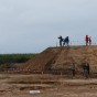 Blick auf die Ausgrabungsfläche, dort sind Wege mit Flatterband gekennzeichnet. Die plane Oberfläche ist im Vordergrund bewachsen, dahinter sind dunkle Flecken. Zahlreiche Besucher befinden sich auf der Fläche, rechts eine Schautafel im Hintergrund ein Maisfeld und eine Aufschüttung, von der Besucher auf die Fläche herunterblicken.