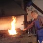 Besucher beobachten den Schmied, der ein Stück Eisen in das lodernde Feuer hält. Darüber befindete sich eine Esse, im Hintergrund hängen Werkzeuge an der Wand, von der Decke hängt eine Werkstattlampe.