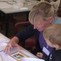 Bild aus dem Kinderzelt. An einem Tisch sitzt eine Kollegin mit einem Pinsel in der Hand, vor ihr liegen Reliefs, Farben und weitere Pinsel. Ein Junge steht neben ihr. 