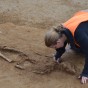 Zwei Personen knien auf dem Boden und legen ein Skelett frei. Zwischen den Fußknochen befindet sich eine Grabbeigabe.