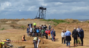 Mehrere Menschen stehen vor mit Flatterband abgesperrten Grabungsflächen, auf der Archäologen ihre Funde freilegen. Im Hintergrund befindet sich ein großer Bagger im Tagebau. Foto: Dirk Bongers