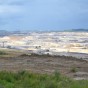 Mehrere Menschen stehen vor mit Flatterband abgesperrten Grabungsflächen, auf der Archäologen ihre Funde freilegen. Im Hintergrund befindet sich ein großer Bagger im Tagebau.