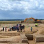 Mehrere Menschen stehen vor mit Flatterband abgesperrten Grabungsflächen, auf der Archäologen ihre Funde freilegen. Im Hintergrund befindet sich ein großer Bagger im Tagebau. Auf der Ausgrabungsfläche ist ein aus Steinen von einem Meter Stärke errichteter massiver Wehrturm mit den Außenmaßen sieben mal sieben Meter gefunden worden, der präsentiert werden konnte.