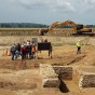 Von einem erhöhten Standpunkt erstreckt sich der Blick über eine archäologische Ausgrabungsstelle. Im Vordergrund befindet sich ein Profil durch einen Graben, dahinter das Rechteck eines Steinturms. Die archäologischen Befunde sind mit Markierspray hervorgehoben worden. Im linken Bereich des Bildes sind mehrere durch Absperrband markierte Wege zu erkennen, auf denen Besucher einzeln und in kleinen Gruppen laufen. Im Hintergrund befindet sich in der Bildmitte ein Mobilbagger, über den Horizont ragen die Schlote und Rauchfahnen des Kohlekraftwerks Weißweiler.