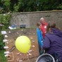 Vor einem Tisch im Freigelände stehen Kinder, dahinter Mitarbeiter und erklären archäologische Funde. Auf einer Stellwand sind Scherben aufgeklebt. Davor steht ein Kinderwagen mit einem gelben Luftballon.