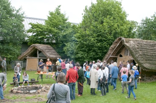 Foto: Zahlreiche Besucher und gehen auf einer Wiese zwischen zwei Lehmgebäuden herum, einige sitzen auf einer Bank am Lagerfeuer.