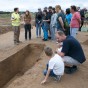 Eine Gruppe Besucher steht vor einer Eintiefung im Boden, ein Archäologe in Warnweste erklärt etwas.
