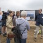 Eine Gruppe Besucher steht vor einem Poster. Ein Mann erklärt etwas, links ein Mann mit Filmkamera.