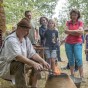 Ein Mann sitzt aneinem kleinen Ofen aus dem Flammen kommen. Besucher sstehen hinter ihm.