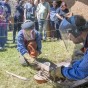 Mehrere Besucher betrachten zwei kniende Personen in Schutzkleidung. Sie gießen flüssige Bronze in eine Gussform, dahinter ein Lehmhaus mit Strohdach.