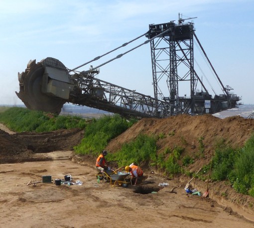 Im Vordergrund arbeiten zwei Studenten mit Schaufel und Spitzhacke an einem Grabungsschnitt. Wenige Meter im Hintergrund nähert sich der Schaufelradbagger der RWE der Grabungsfläche.