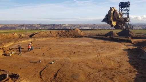 Das Foto zeigt einen Kreisgraben als Verfärbung im Boden mit drei Archäologen und den Schaufelradbagger im Hintergrund. 