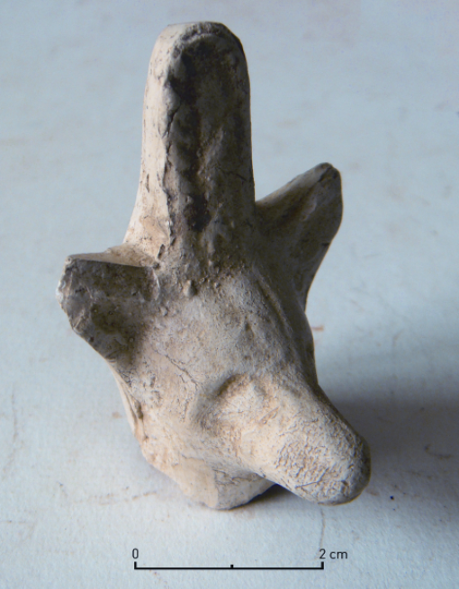 Abgebrochener Oberteil einer kleinen Statuette aus weißem Ton. Das auf den ersten Blick an einen Hund oder Fuchs erinnernde Kopf weist zwischen den Ohren einen zungenförmigen Fortsatz auf, der ihn in eine Reihe römisch-ägyptischer Götterstatuetten stellt und eine Deutung als Darstellung des Gottes Anubis erlaubt.