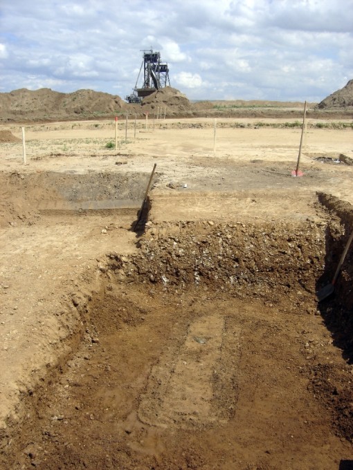 Das Foto zeigt im Vordergrund ein spätantikes Körpergrab mit einer Glasschale, die umgedreht ungefähr in der Mitte des Grabes liegt. Im Hintergrund ist ein Braunkohlebagger zu sehen.