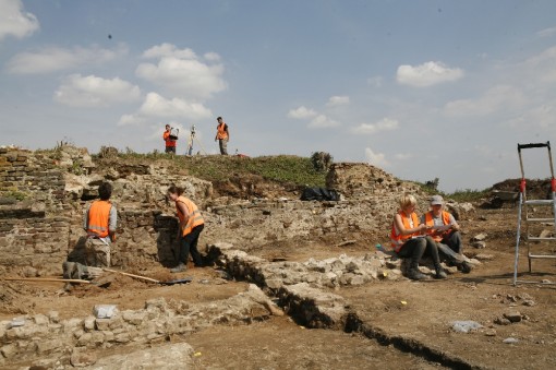 Das Foto zeigt eine Grabungssituation, bei der mehrere Archäologen zwischen Mauerfundamenten arbeiten. Im Hintergrund befindet sich ein Hügel, auf dem ein Vermessungsgerät steht.