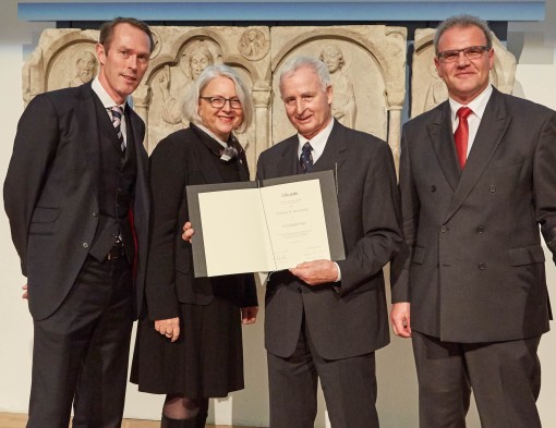 Der Preisträger hält die Urkunde in der Hand und wird umrahmt von einer Dame und zwei Herren.