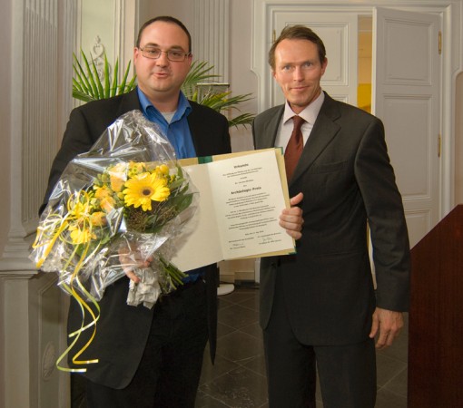 Ein Herr hält einen Blumenstrauß und eine Urkunde in den Händen. Daneben steht ein weiterer Herr.