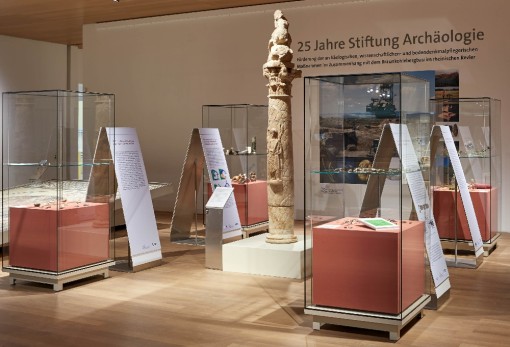 Vor einer Wand mit der Aufschrift 25 Jahre Stiftung Archäologie befinden sich vier Vitrinen, mit Fundstücken aus dem rheinischen Braunkohlenrevier. Neben den Vitrinen stehen Aufsteller mit textlichen Erläuterungen.